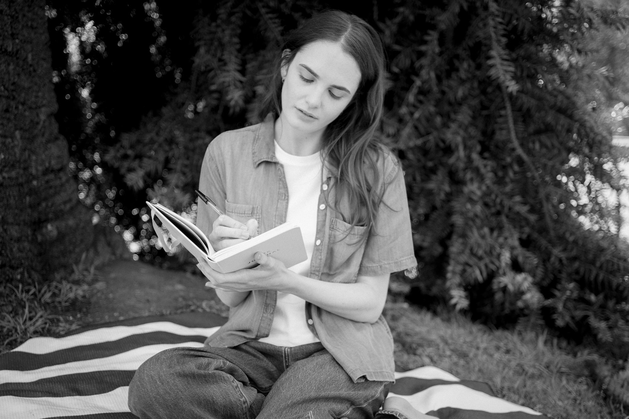 A black and white photo of Tayla sitting outside on a picnic mat against a tree. She is writing in a notebook and wearing a short-sleeved denim shirt, white t-shirt and jeans.