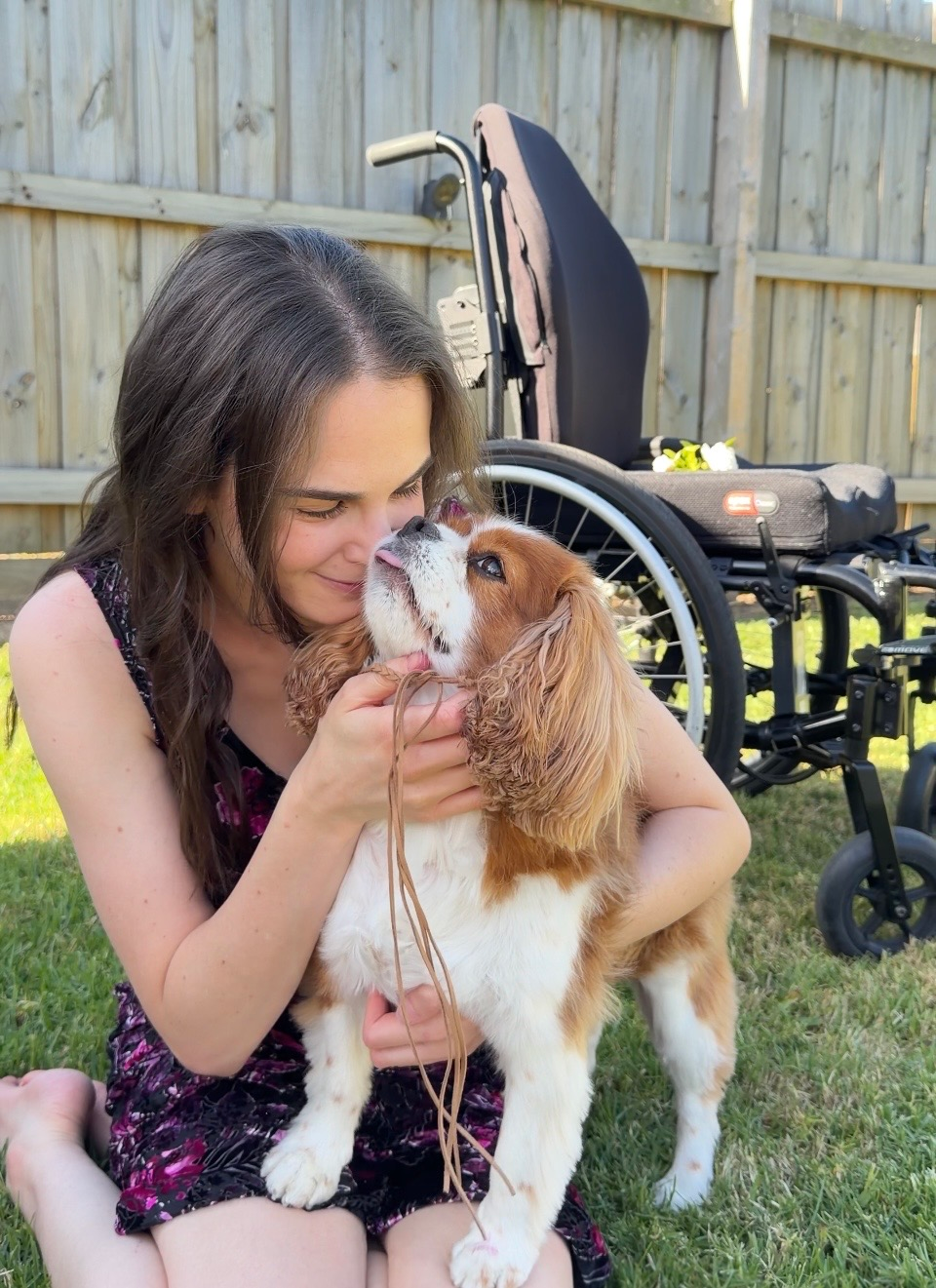 Tayla (wavy brown hair, wearing a black and purple midi dress) is hugging her Dog Cleo (a ruby and white King Charles Cavalier Spaniel) they are sitting on grass outside with Tayla's black manual wheelchair in the background with a white rose on it.