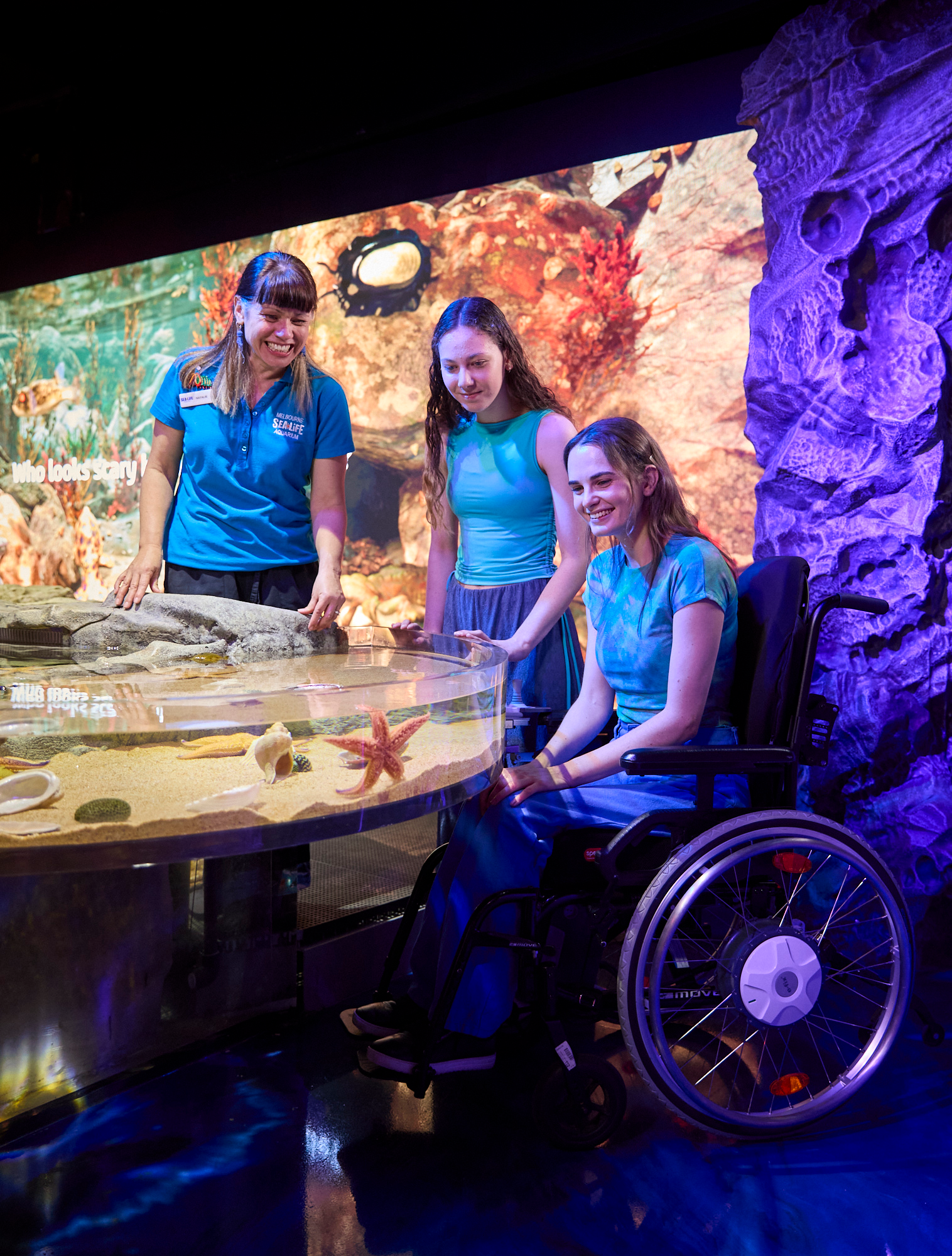 Tayla sits in her wheelchair at an accessible rock pool with her younger (early twenties) sister standing next to her and a staff member from the Melbourne Aquarium. All three women are wearing blue tops and looking at a star fish on the side of the transparent rock pool.