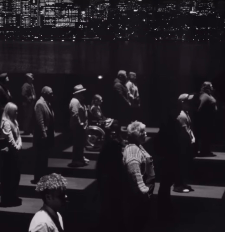 A black and white image of dozens of people standing in row on squares of lights or in shadows. Tayla is amongst them in her wheelchair.