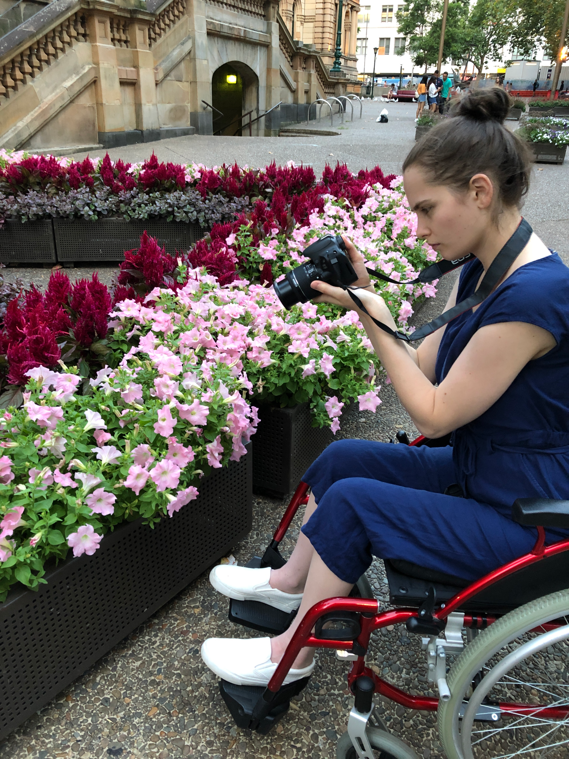 Tayla (wearing a blue jumpsuit, brown hair in a loose bun) sits in her red wheelchair outside taking pictures with a DSL camera of red and pink flowers in a flower box arrangement.