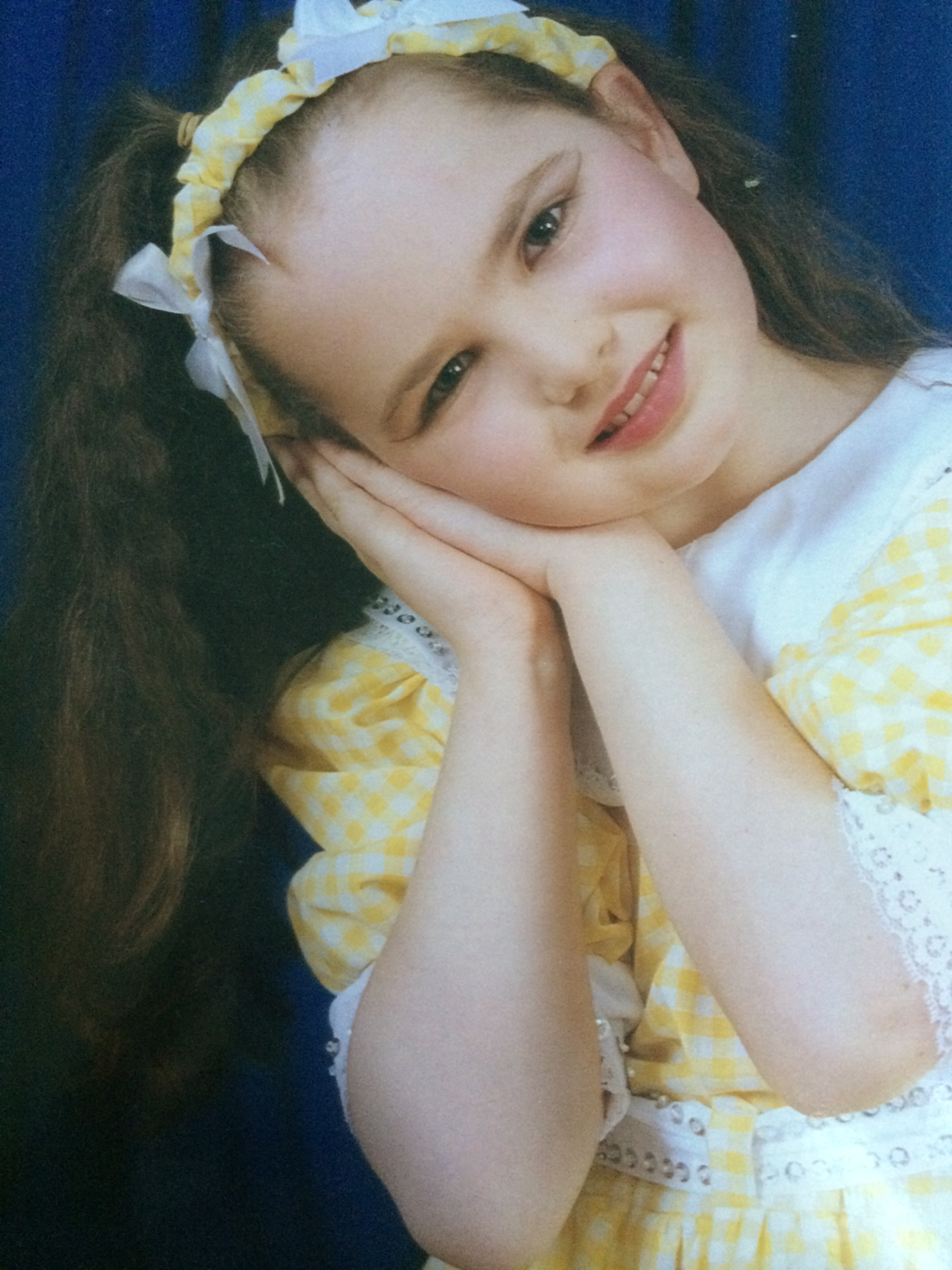 Posed dancing picture close up of Tayla as a child (6 years old). She is smiling, has long brown curl hair, dance make up and a white and yellow checked dress and headband.