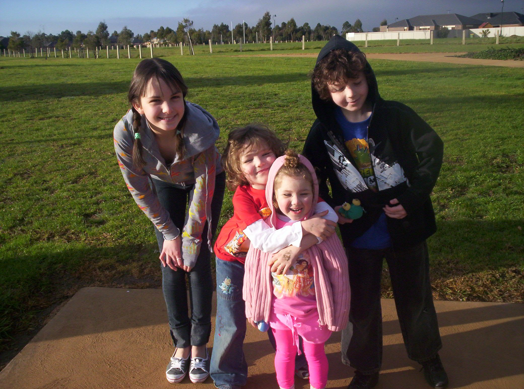 15 year old Tayla (Brown hair with fringe and two plaits, wearing a grey hoodie with stars and jeans) is standing crouched next to her three younger siblings: toddler sister (with brown curly hair, wearing a pink hoodie and pants), 6 year old brother (with dark blond curly hair, wearing a red hoodie and jeans) hugging little sister, and 8 year old brother (wearing black hoodie and jeans) leaning in. All are standing outside and smiling to camera.