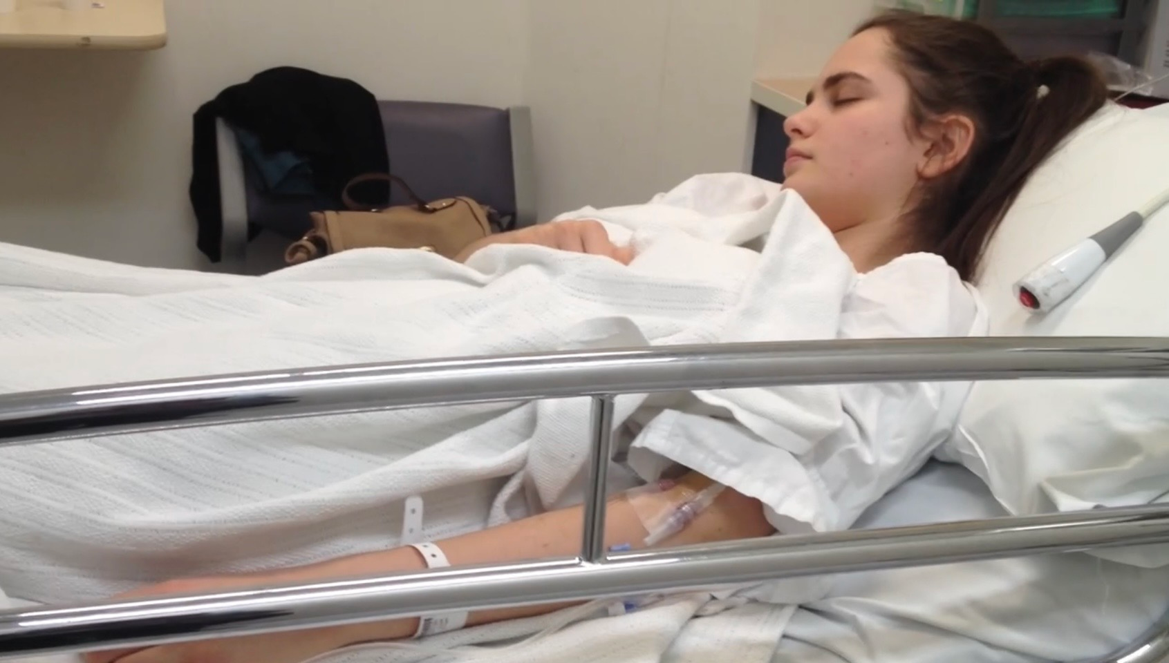 Tayla lays in a hospital bed, with silver rails raised and is wearing a white hospital gown and wristband.