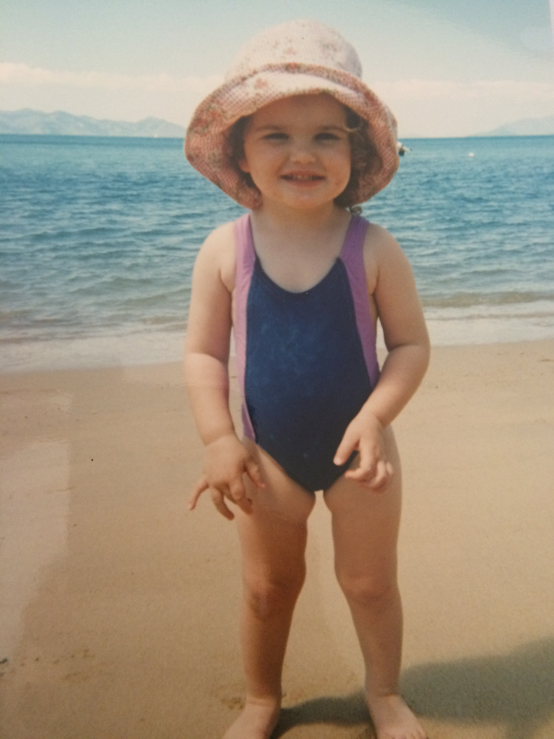 Tayla as a toddler (2 years old) on sand at a beach in front of of blue still water. She is smiling and wearing a pink sun hat and blue and purple one piece bathers.
