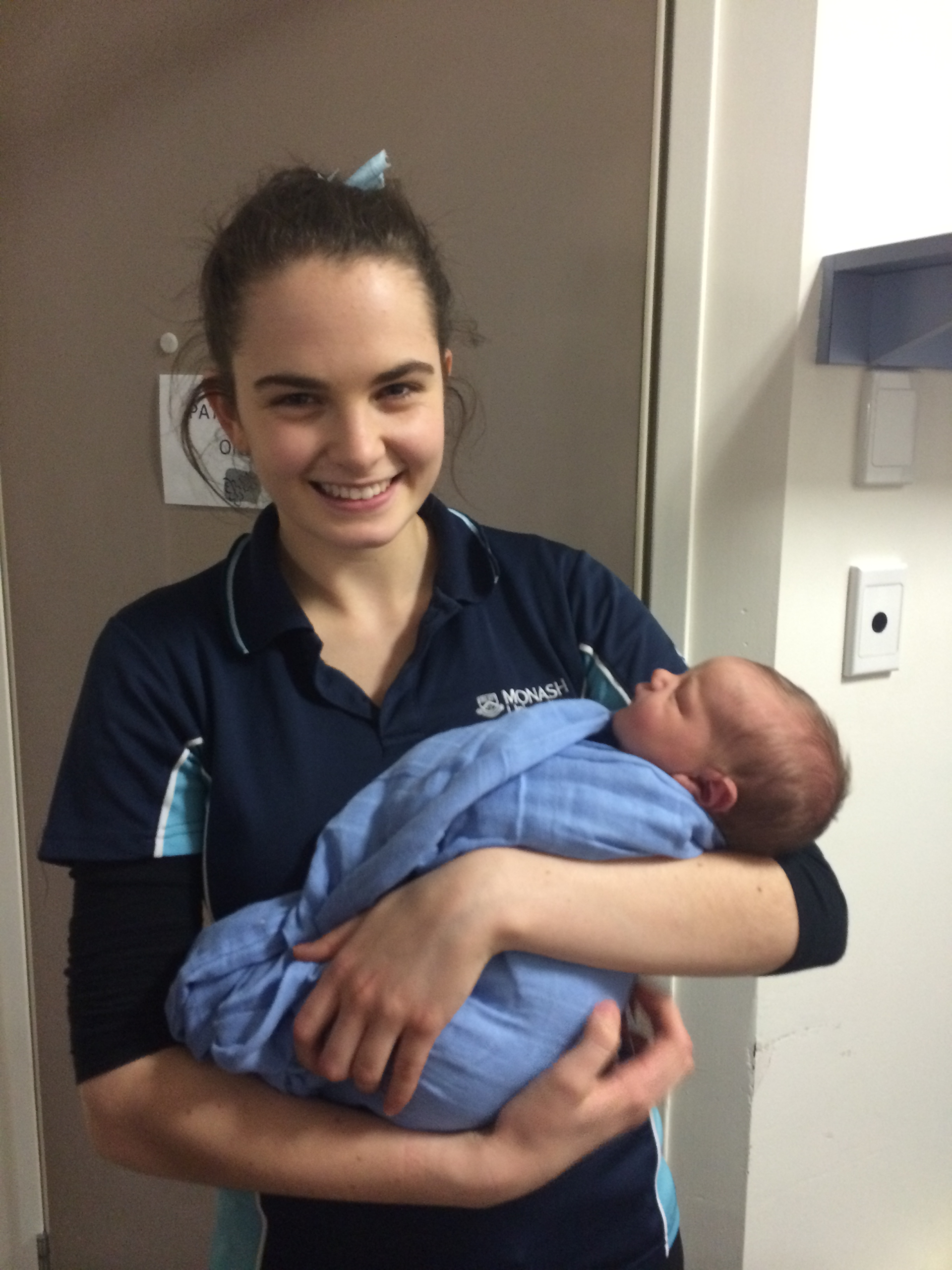 Tayla at a hospital, in a university uniform and bun is holding a newborn baby wrapped in a blue blanket. She is smiling to camera.