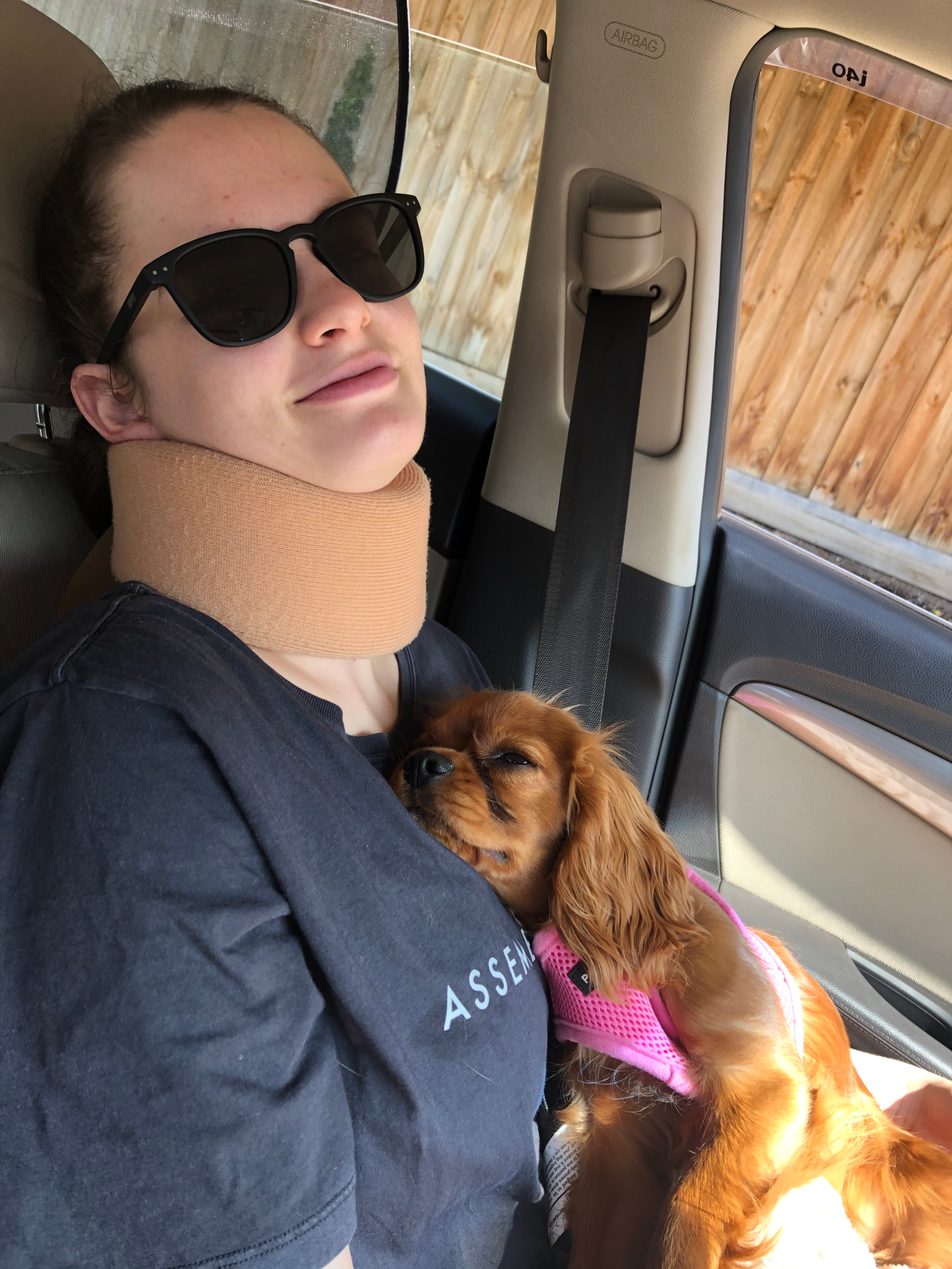 Tayla is in the car laying against the seat with a tan neck brace on and sunglasses. She holds puppy Mia (a small ruby King Charles Cavalier Spaniel) who is wearing a pink harness and laying against Tayla's chest.