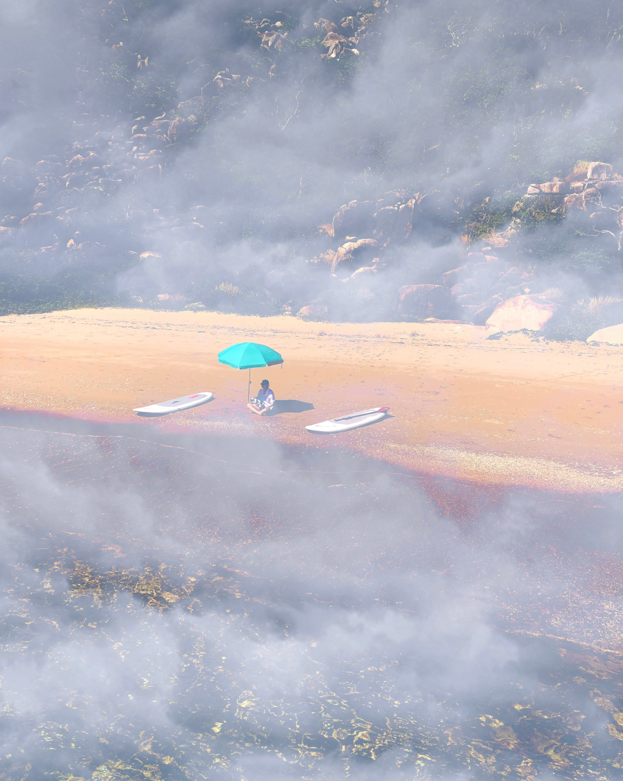 A very smoky scene on a beach with a figure sitting on the sand under a small umbrella with two paddle boards,