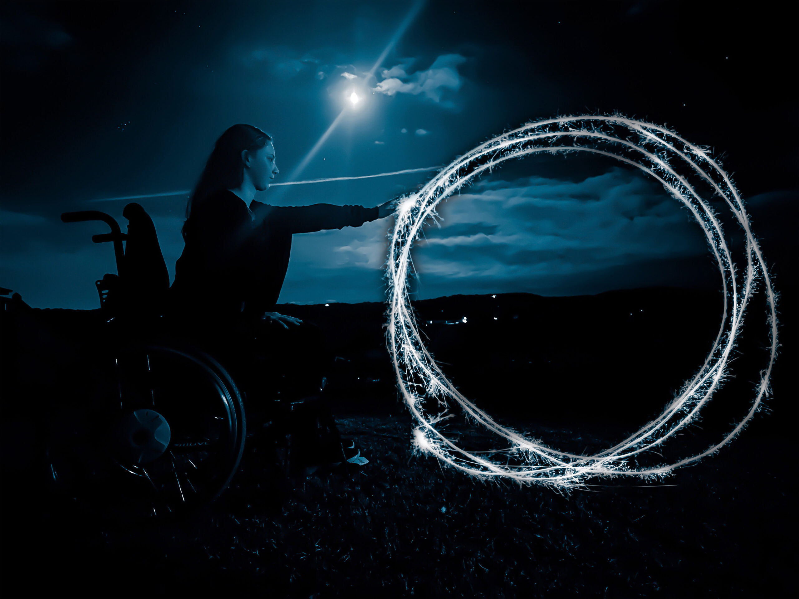 a dark slate blue and black scene with a moon shinning over a profile view of a feminine figure sitting in a wheelchair touching large rings of light/sparks.